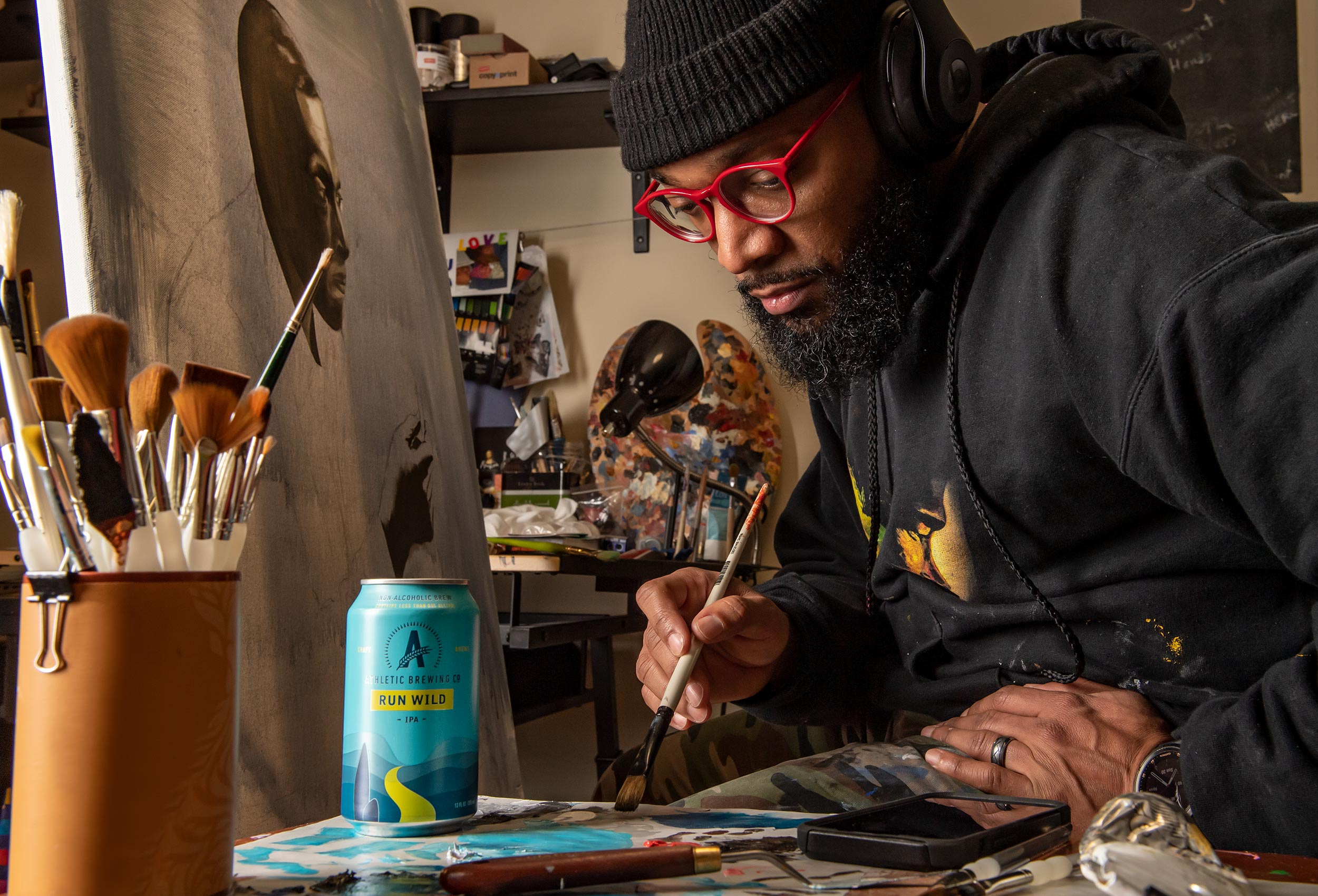 Athletic Brewing Co. Ambassador M. Anthony Cooper-Jenkins paints in studio  with can of Run Wild non-alcoholic beer