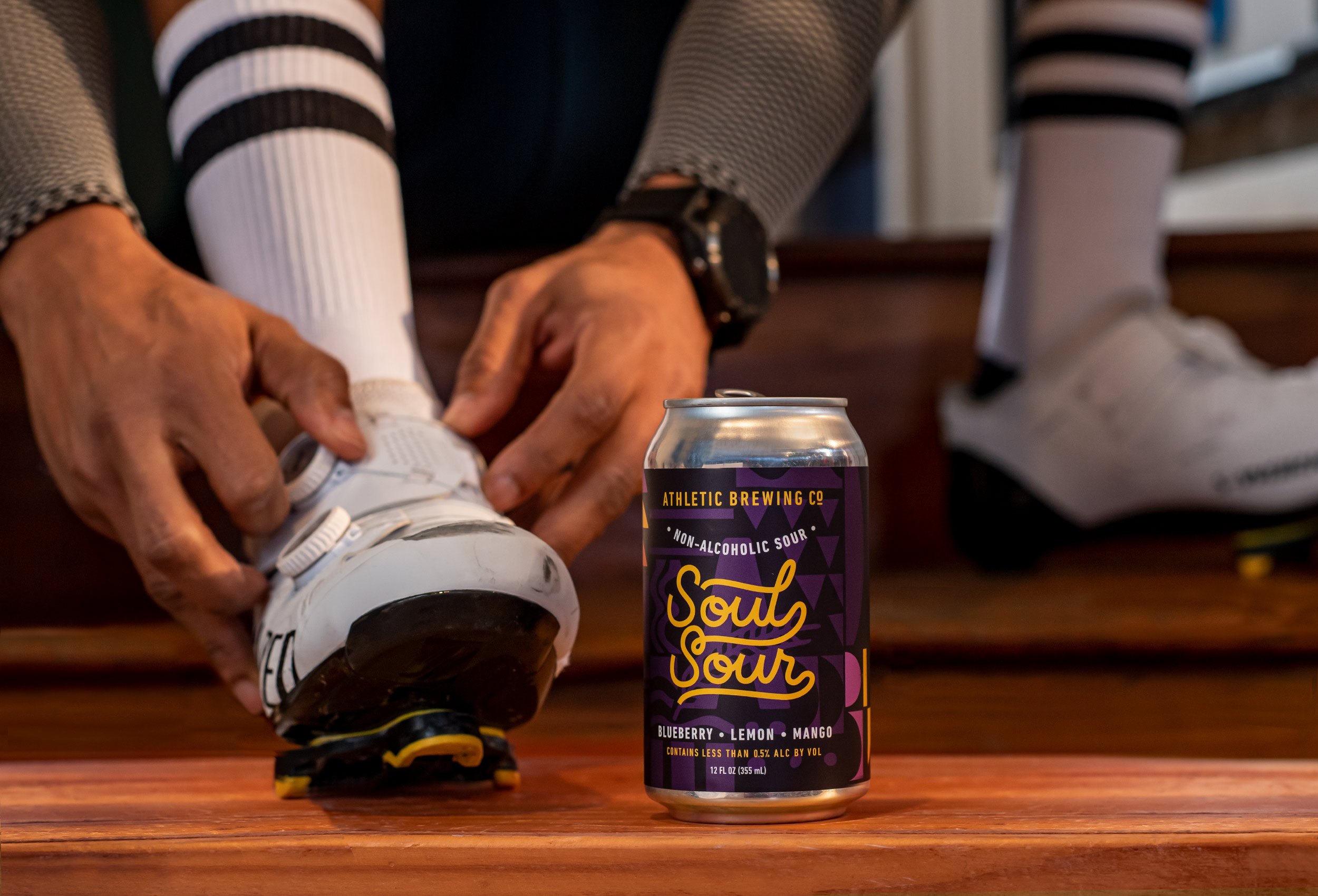 Cyclist laces bike shoes on wood floor with can of Soul Sour non-alcoholic beer