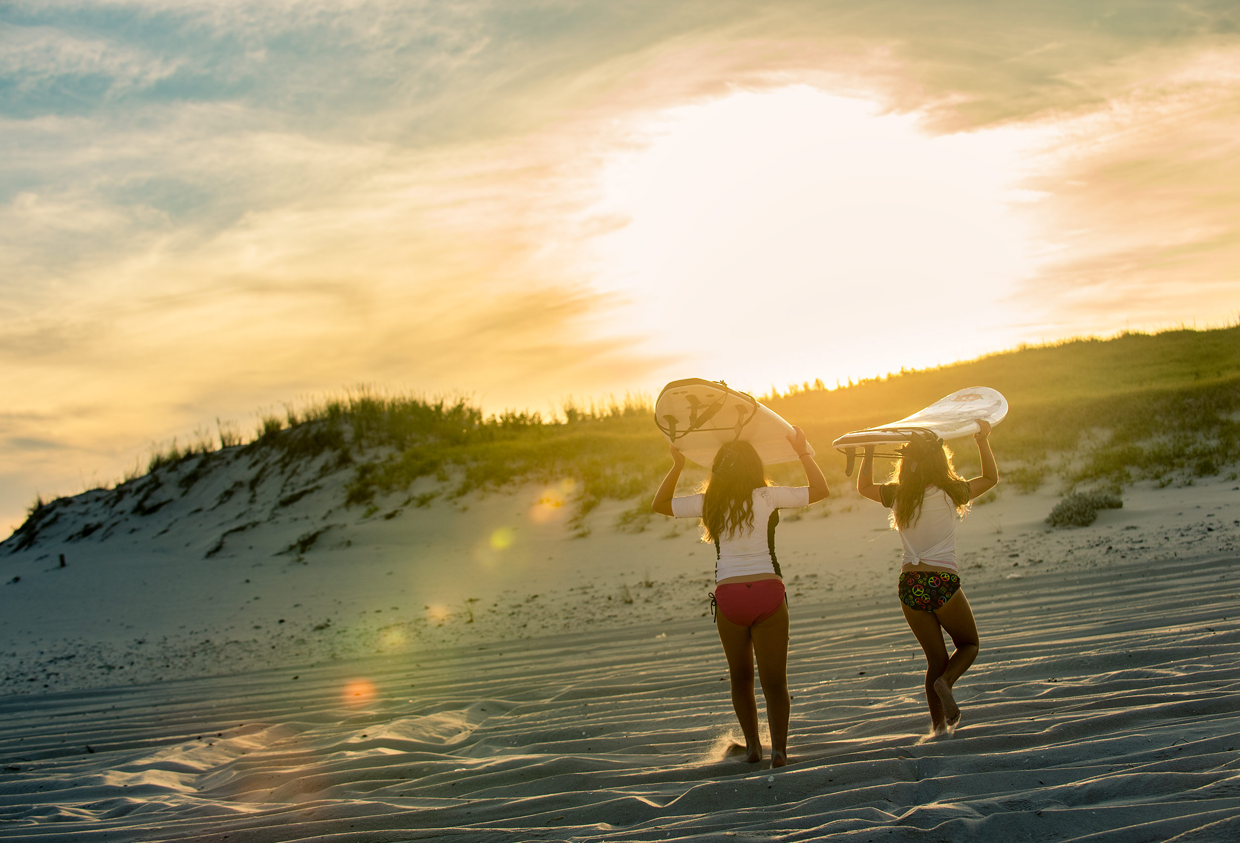Surfer girls walk towards dunes with surfboards on beach at sunset