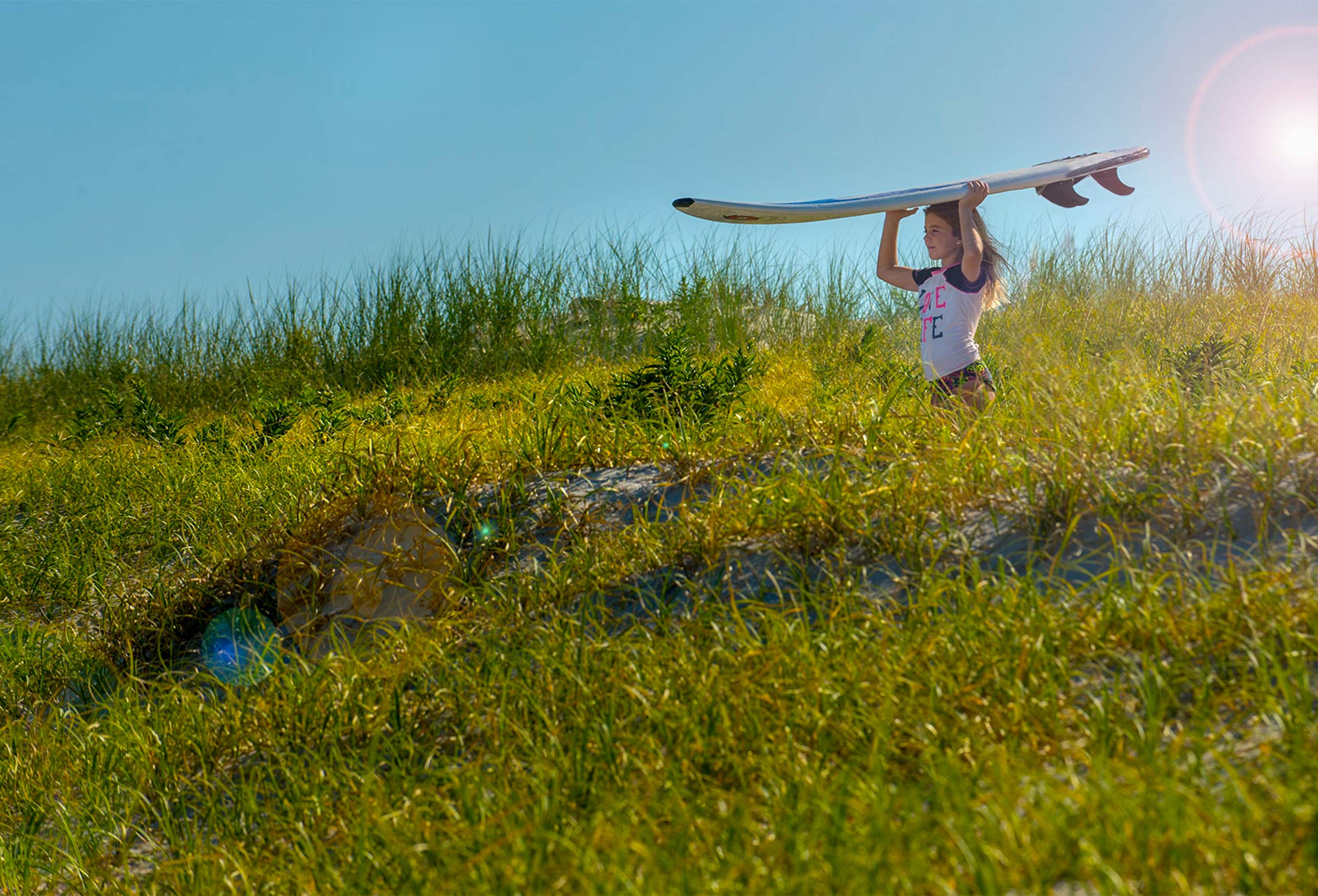 Teen surfer girl walks through dunes with surfboard on head